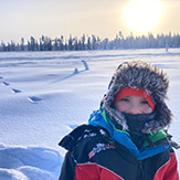 Kind im Schnee, ganz vermummt, im Hintergrund Wintersonne.