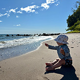 Baby am Strand, sitzt im Sand, Meer und Steilküste.