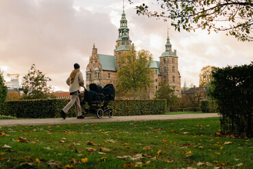 Eine Person spaziert mit einem Kinderwagen durch einen Park. Im Hintergrund ist ein Schloss zu sehen.