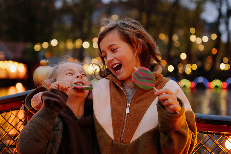 Zwei Kinder essen jeweils einen bunten Lolli vor einer beleuchteten Stadt.