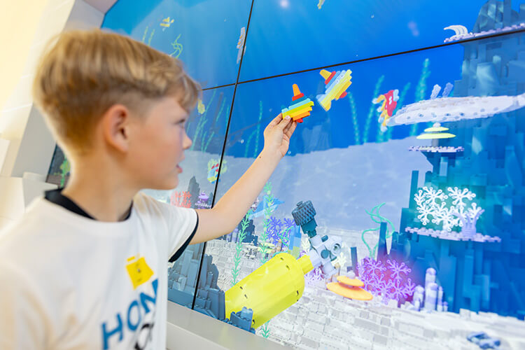 Junge hält zwei kleine bunte Lego-Fische vor eine große digitale Unterwasserwelt mit Korallen und Fischen.