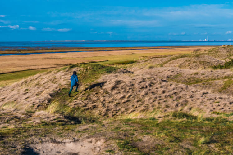 Kind in blauer Jacke läuft über bewachsene Dünen mit Blick auf das Meer und einen blauen Himmel.