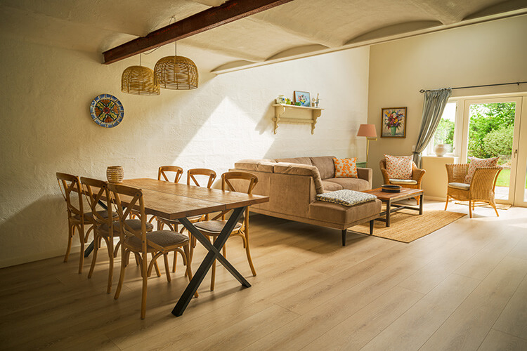 Helles Wohnzimmer mit Holztisch und Stühlen, Sofa, Sessel, Teppich und großen Fenstern mit Blick ins Grüne.