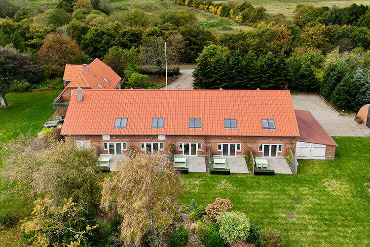 Luftaufnahme eines langgestreckten Hauses mit roten Dachziegeln und mehreren Terrassen, umgeben von Rasen und Bäumen.