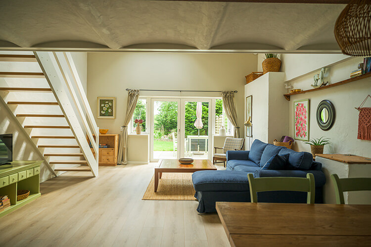 Helles Wohnzimmer mit blauer Eckcouch, Holztisch, Treppe links und großen Fenstertüren zum Garten.