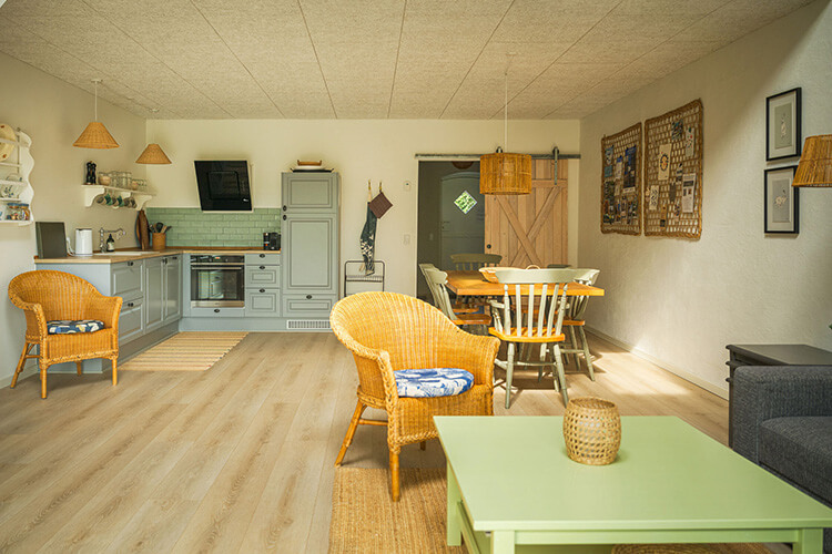 Wohnzimmer mit offener Küche, zwei Korbsesseln, Esstisch mit Stühlen, Sofa und Wanddekorationen.