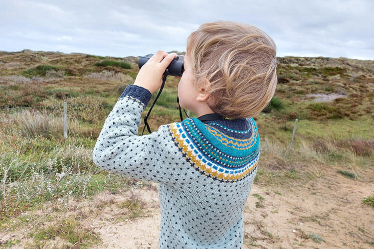 Kind mit hellblondem Haar und blau-grauem Pullover blickt durch ein Fernglas in eine Heidelandschaft.