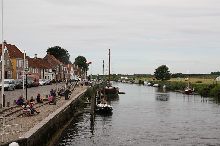 Fluss mit angelegten Segelbooten und Uferpromenade mit Menschen vor Reihenhäusern unter bewölktem Himmel.