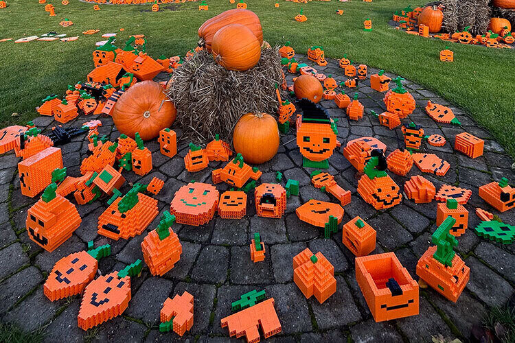 Mehrere orangefarbene Kürbisse und zahlreiche aus Bausteinen zusammengesetzte Kürbisfiguren mit Gesichtern auf einem gepflasterten Kreis und einer Wiese im Hintergrund.