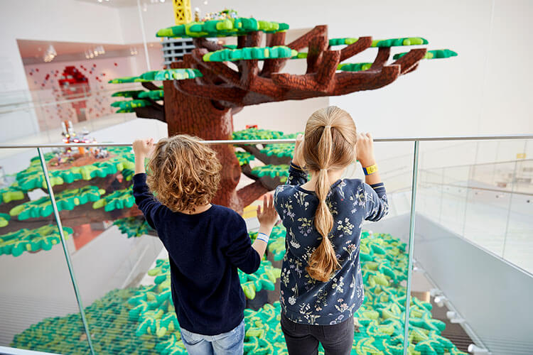 Zwei Kinder stehen vor einem großen Baum aus Legosteinen und schauen hindurch auf die Ausstellung.