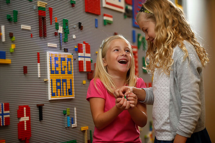 Zwei Mädchen vor einer Wand mit bunten LEGO-Steinen, eines lacht und hält LEGO-Steine in den Händen.