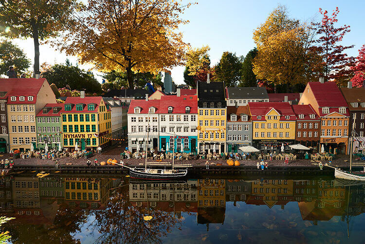 Reihe bunter Häuser aus Legosteinen am Wasser mit Booten und herbstlich gefärbten Bäumen im Hintergrund.