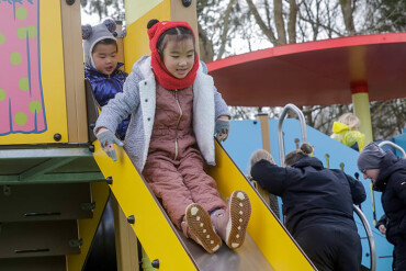 Zwei Kinder rutschen auf einer gelben Rutsche auf einem Spielplatz, im Hintergrund stehen mehrere Erwachsene.