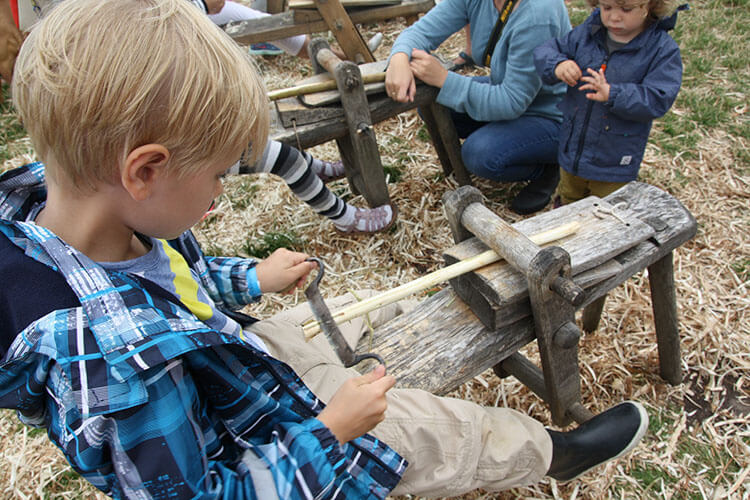 Kind sitzt auf einer Holzbank und schnitzt mit einem Messer an einem Holzstück, im Hintergrund weitere Kinder auf dem Boden.