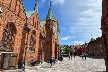 Backsteinkirche mit spitzen Türmen an einem gepflasterten Platz in einer historischen Stadt mit weiteren Fachwerkhäusern im Hintergrund.