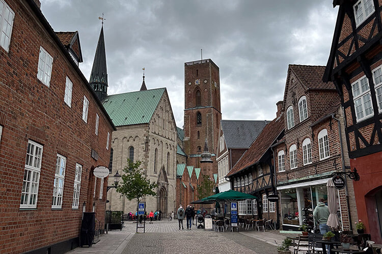 Gepflasterte Straße mit Fachwerkhäusern und einer Kirche mit Turm unter bewölktem Himmel.