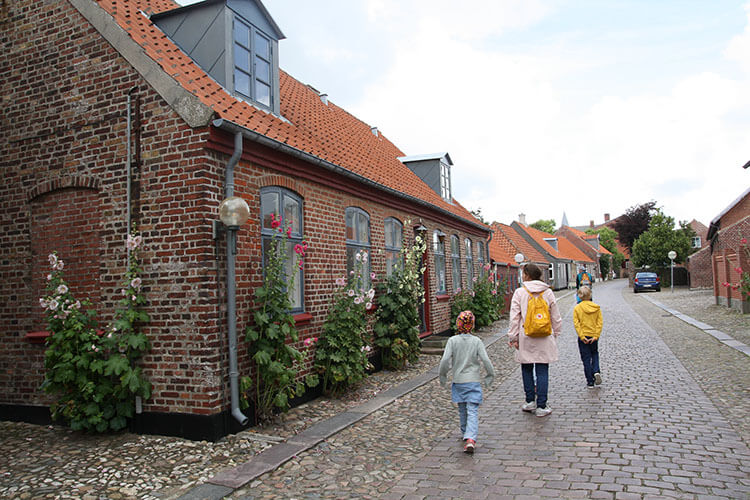 Drei Personen mit Rucksäcken gehen eine gepflasterte Straße entlang an einem Backsteinhaus mit roten Ziegeldächern und Rosensträuchern.
