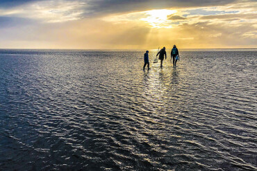 Drei Personen gehen bei Sonnenuntergang durch flaches Wasser mit weitem Horizont.