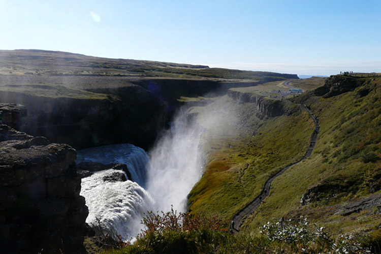 Wasserfall in einer felsigen Schlucht mit Nebel und einem Pfad am rechten Hang unter klarem Himmel
