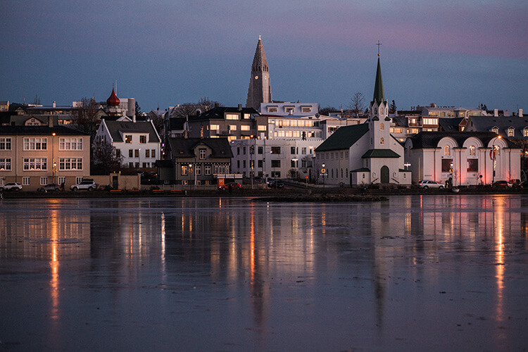 Abendliche Stadtansicht von Reykjavik mit beleuchteten Häusern, zwei Kirchen mit spitzen Türmen und spiegelnder Wasseroberfläche im Vordergrund.
