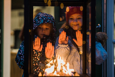 Zwei Kinder mit Winterkleidung schauen durch eine Glasscheibe auf ein offenes Feuer und halten die Hände zum Wärmen davor