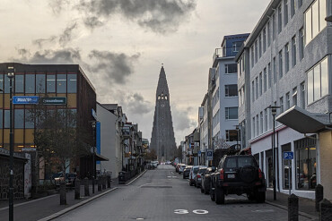 Blick auf die Hallgrímskirkja vom Ende einer Straße mit parkenden Autos und mehrstöckigen Gebäuden auf beiden Seiten bei bewölktem Himmel