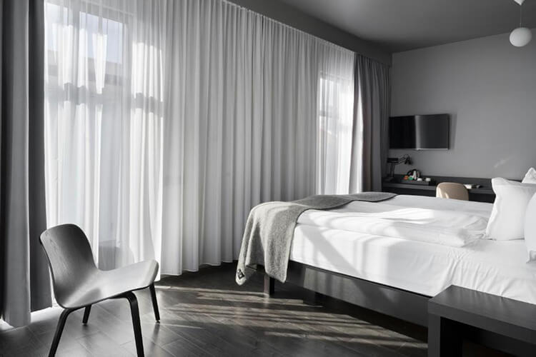 Modern eingerichtetes Hotelzimmer mit großem Bett, Stuhl vor bodentiefen Fenstern und Fernseher an der Wand.