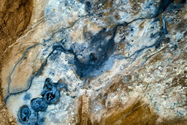 Luftaufnahme einer geothermalen Landschaft mit bläulichen Wasserstellen und einem Holzsteg in der Mitte