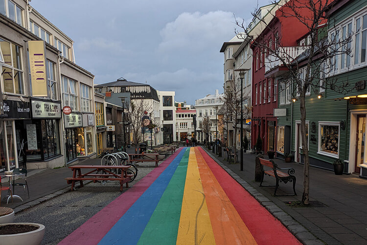 Straße mit breitem Regenbogenstreifen in der Mitte, umgeben von mehrstöckigen Gebäuden und Bänken auf beiden Seiten.