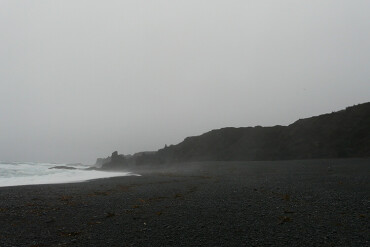 Nebelverhangener schwarzer Strand mit dunklen Felsen im Hintergrund und welligem Meer