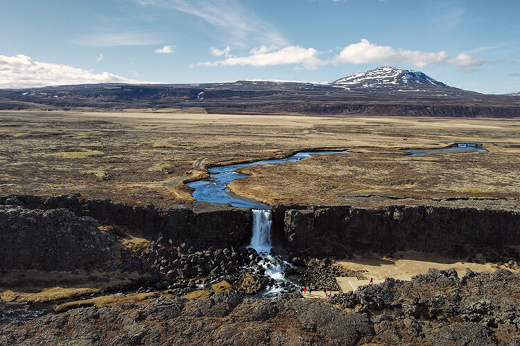 Landschaft mit Fluss, der über einen Wasserfall in eine Felsschlucht stürzt, im Hintergrund ein schneebedeckter Berg.