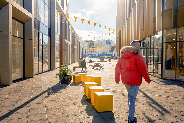Person mit roter Winterjacke und Jeans geht durch eine von modernen Gebäuden gesäumte Fußgängerzone mit gelben Sitzwürfeln und Wimpelketten.