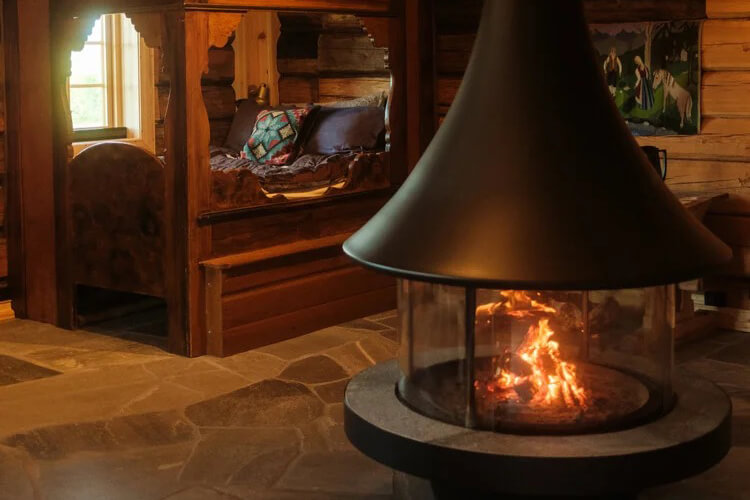 Im Vordergrund ist ein Kamin mit einem Feuer zu sehen, im Hintergrund steht ein antikes Holzbett.