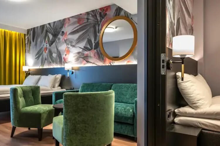 Zwei Betten in einem Hotelzimmer, die von einer Wand voneinander abgetrennt sind. In der Mitte steht eine grüne Sitzgruppe.