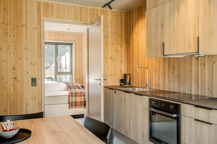 Kleine Küche mit Holzvertäfelung, Einbauherd, Spüle, Wasserkocher und Blick in ein Schlafzimmer mit Bett und kariertem Plaid