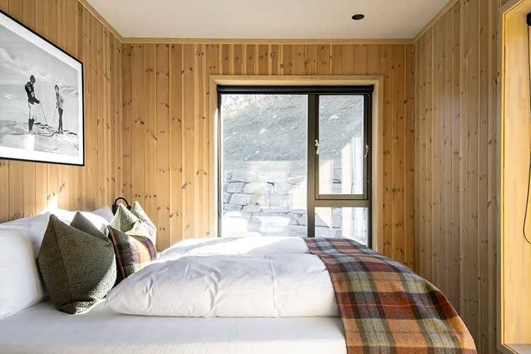 Schlafzimmer mit Holzwandverkleidung, großem Bett mit weißen Decken, kariertem Überwurf und mehreren Kissen sowie einem Fenster mit Blick auf eine Steinmauer