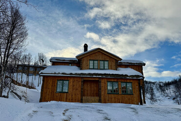 Holzferienhaus mit grün gerahmten Fenstern und Schnee auf dem Dach vor winterlicher Landschaft in Norwegen