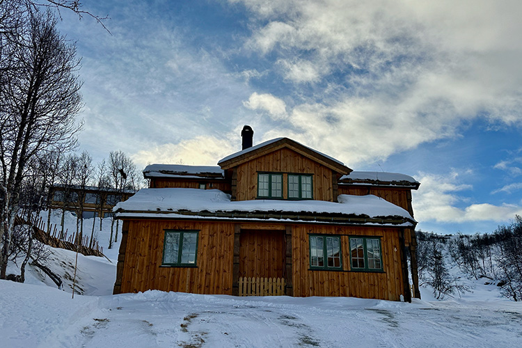 Holzferienhaus mit grün gerahmten Fenstern und Schnee auf dem Dach vor winterlicher Landschaft in Norwegen