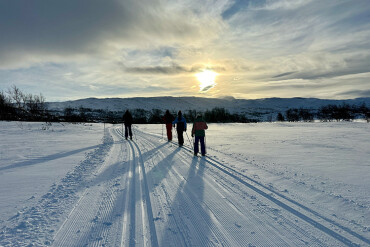 Gruppe von Langläufern auf einer präparierten Spur in einer verschneiten Winterlandschaft mit Bergen im Hintergrund und tiefstehender Sonne am bewölkten Himmel