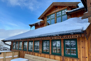 Holzgebäude mit grün gerahmten Fenstern und schneebedecktem Dach vor blauem Himmel
