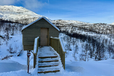 Kleines Holzhaus mit Sauna drin. Treppe und Geländer in verschneiter Winterlandschaft vor schneebedeckten Bergen.