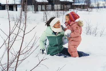 Frau in grüner Winterjacke und Kind in rosa Schneeanzug mit bunter Mütze sitzen lachend im Schnee vor Häusern