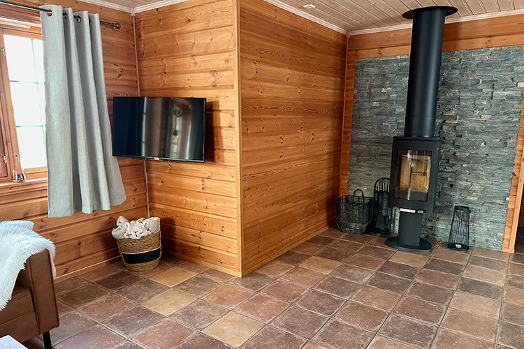 Wohnzimmer mit Holzverkleidung, Kaminofen vor Steinwand, Wandfernseher und Korb mit Decken neben Fenster mit Vorhang