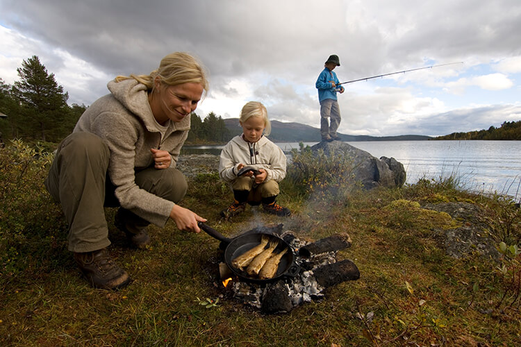 Eine Frau und ein Kind braten auf einem Lagerfeuer Fische, im Hintergrund steht eine Person mit einer Angel.