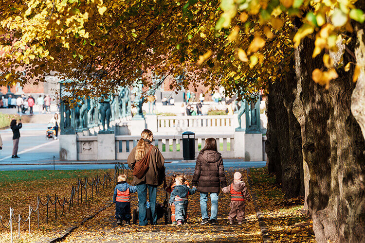 Zwei Personen laufen mit drei Kindern an den Händen durch einen Park, die Bäume haben buntes Laub.
