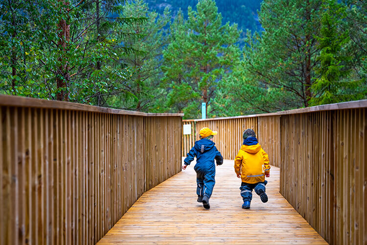 Zwei kleine Kinder rennen in Regenkleidung mit dem Rücken zur Kamera auf einer Holzbrücke.