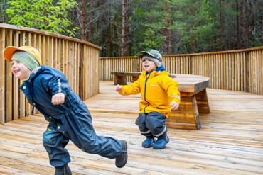 Zwei kleine Kinder rennen in Regenklamotten über einen Holzpfad.