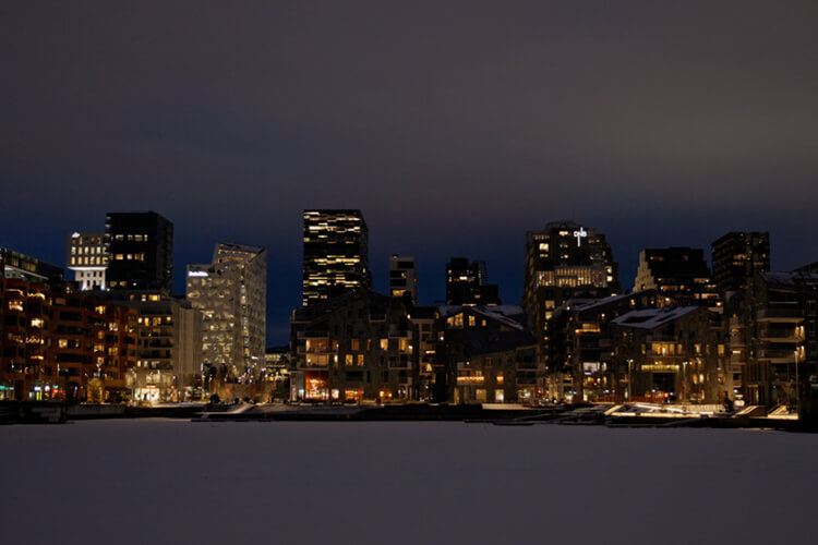 Blick auf Wasser und auf hohe Gebäude in Oslo bei Nacht. In den Fenstern der Gebäude brennt Licht.