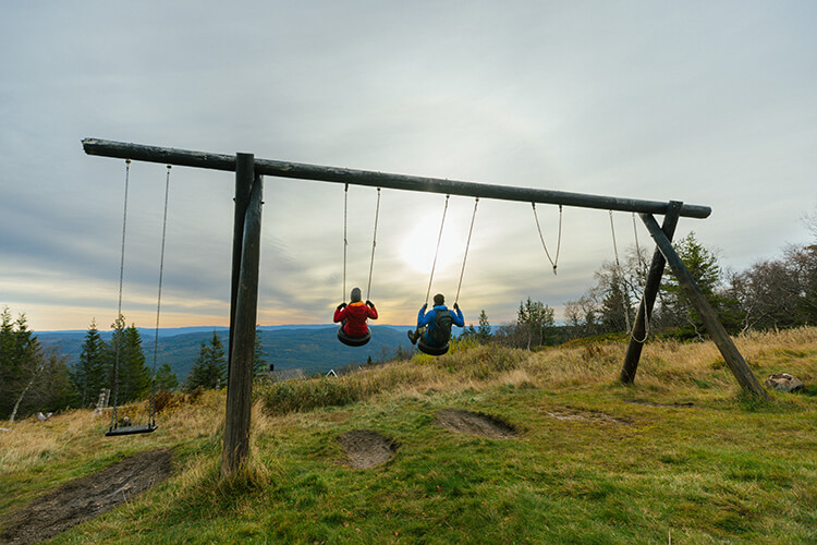 Zwei Personen sitzen auf einer großen Schaukel mit Blick in eine hügelige Landschaft.