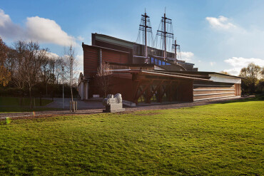 Das Vasa-Museum in Stockholm zeigt ein großes hölzernes Schiff, das teilweise aus dem Museumsgebäude herausragt, umgeben von einer grünen Wiese und Bäumen unter blauem Himmel mit einigen Wolken.